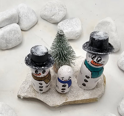 Schneemänner Miniatur, gefertigt aus  kleinen Holzkegeln, auf Sandstein mit Bäumchen.