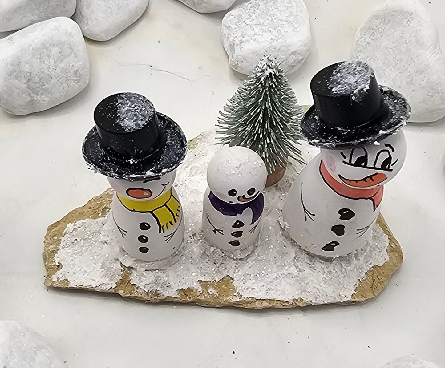 Schneemänner Miniatur, gefertigt aus  kleinen Holzkegeln, auf Sandstein mit Bäumchen.